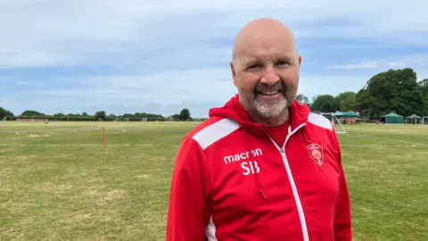 A bald man with a greying beard, wearing a red AFC Corsham tracksuit and looking into the camera. Behind him a playing field with white lines drawn out on the grass, green gazebos and flags. 