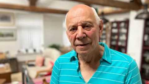 Roy Barnett - elder man with white hair, bald patch in the middle with a slight smile and furrowed brow, wearing an aqua blue collared shirt with thin white stripes, blurred background of a living room