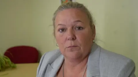 Jamie Niblock/BBC Collette Kerry wears a light grey jacket with a silver necklace hanging around her neck. She has tied-back blonde hair and is looking at the camera. She is sitting in a room with a red chair and a table in the background.