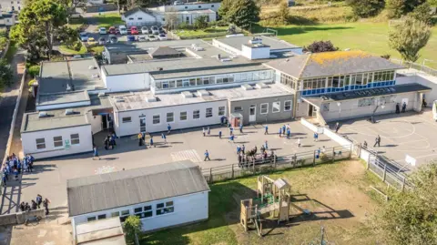 States of Guernsey A drone shot of St Anne's School on a sunny day. The playground has lots of pupils wearing blue uniforms. There are a series of connected buildings and a football pitch beyond and wooden play castle in the foreground. 