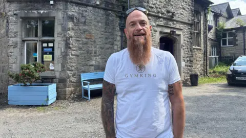 James Deakin stands outside historic large grey brick building with a light blue bench in front of it. He is bald with a long beard with sunglasses on his head and has a sleeve of tattoos.