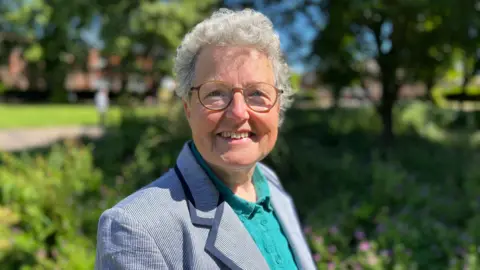 Jo Thewlis/BBC Catherine Rowett is wearing a light blue blazer over an aquamarine shirt. She's standing outside with a leafy background behind her.