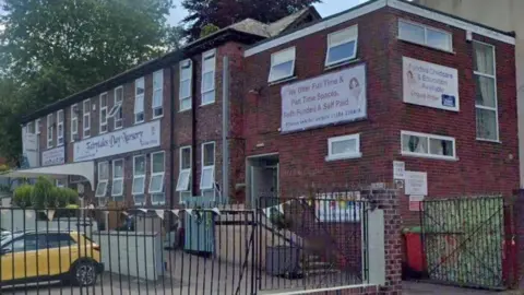 Google A red brick two-storey building behind metal railings, with a sign reading 'Fairytales Day Nursery'. Banners advertise funded childcare and full and part-time spaces. A yellow car is parked outside.