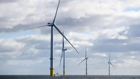 Four wind turbines tower over the sea. There are white clouds and some blue sky above.