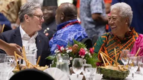 UK Government UK Prime Minister Sir Keir Starmer has short greying hair and weard brown thin-rimmed glasses with a blue suit over a white shirt. He chats to Samoa Prime Minister Fiamē Naomi Mataʻafa, who has short grey hair and wears a green, orange and pink outfit, as they sit at a table decorated with flowers and laden with several wine glasses.