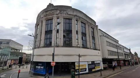 The frontage of a curved building with high double windows set between concrete pillars. On the top of the building the name Beatties can be seen. The building is clearly closed with hoardings around its ground floor