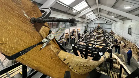 A view of the longship in the process of being built inside a large warehouse. Volunteers can be seen working on the ship body. Planks of wood lie either side of the ship.