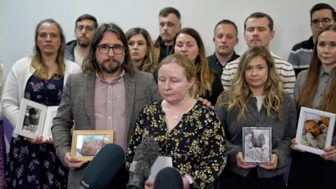 A group of people stand together in front of press microphones, holding framed photographs of babies. Two people stand at the front, one holding a photo, while others stand behind in rows in a plain indoor setting.