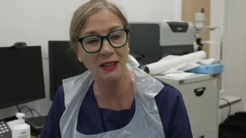 The picture shows a person wearing glasses and a plastic apron, seated in an office or medical environment. Behind them are two computer monitors and various office supplies. Some rolled-up papers rest on a filing cabinet in the background.