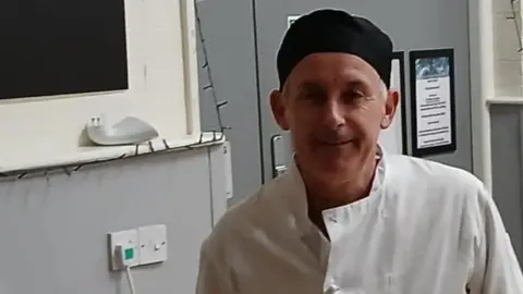 Paul Cleasby smiles at the camera while wearing a chef uniform. He wear a black hat and a white chef's apron. 