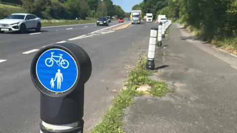 George Carden/BBC A cycle lane has two shades of concrete laid at different times. In the foreground is a sign showing shared use for cyclists and pedestrians and in the background is traffic driving on the A27 near a layby.