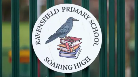 A close-up view of the Ravensfield Primary School sign on dark green railings. The sign is circular and features a drawn picture of a raven bird sitting on a pile of three books on a white background. Around the edge of the circle are the words "Ravensfield Primary School" at the top and "Soaring high' at the bottom.