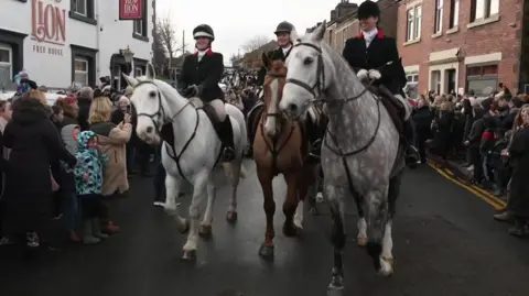 Riders with their hard hats and hunting outfits from The Holcombe Harriers hunt ride through the streets of Wheelton, Lancashire opposdite the RTed Lion Inn.