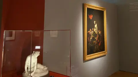 A sculpture of a woman looking towards the floor in a glass case is pictured to the side of a Joseph Wright painting which is lit up.