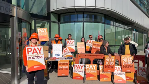BBC More than a dozen banners, with some including the words pay restoration for doctors, are either being held by people outside a building or are vertical on the ground.