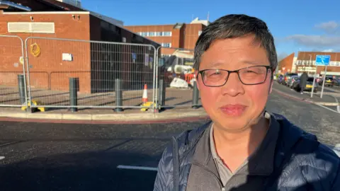 A man, wearing glasses and a blue anorak, looks directly at the camera. Behind him is a hospital car park.