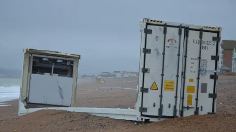 A white container stuck in sand on a beach. The top and sides of the container are missing. The water appears rough and the sky is grey and stormy.