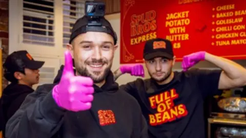 We Create Popular/PA Wire Jacob (left) and Harley Nelson in the kitchen in front of a red menu. Both are wearing black baseball hats and tops. Jacob is doing a thumbs up gesture while wearing a pink latex glove and Harley is in the background flexing his muscles