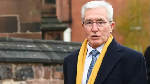 A man with white hair, wearing frameless glasses. He is also wearing a yellow scarf on top of a navy blue coat. He is wearing a blue pattern tie against a white shirt. He stands before a stone wall of red and grey, with grey tiles on the top of it
