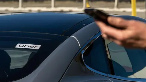 A male hand holding a mobile phone in the foreground with the back of a black car behind him. In the rear window of the car there is an Uber sign.