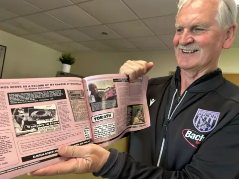 Norman Bartlam Alistair Robertson is wearing a blue West Bromwich Albion training top with the club's badge on and is holding a copy of the book, printed on pink paper, towards the camera