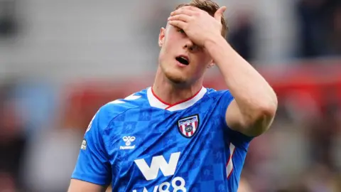 Sunderland's Daniel Ballard holds his forehead, wearing his team's away shirt, which is blue with a repeated pattern depicting a lighthouse through a circular structure