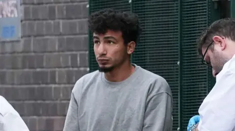 A man with dark hair and a beard is wearing a grey jumper. He is being held by custody officers.