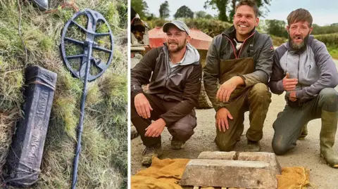 Three men are crouched down on the ground on what appears to be a road. They are smiling with the two ingots placed on the ground in front of them on a mustard-coloured piece of cloth. An agricultural vehicle with a sheepdog perched on it can be seen behind them.