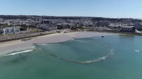 An aerial shot of the Victoria Marine Lake in St Helier. A sandy beach is behind a water-filled the pool, with St Helier behind that. 