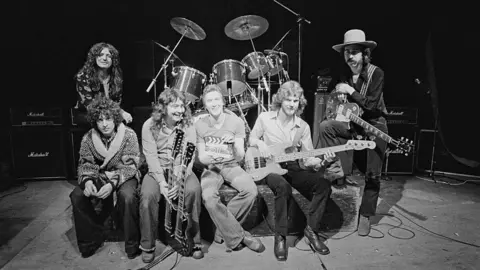 Fin Costello/Redferns/Getty Images A black and white photograph of six men, mainly holding musical instruments, sitting in front of a drum kit.