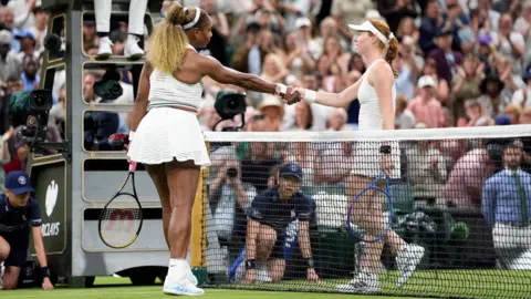 PA Media. Serena Williams y Maya Joint se dan la mano por encima de la red de tenis frente a la silla del árbitro, los recogepelotas y una gran multitud en la tribuna. Ambas visten ropa de tenis completamente blanca.