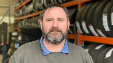 BBC A dark-haired man with a greying beard, wearing a grey jumper over a blue Polo top, standing in front of a tall orange rack of new tyres