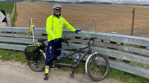 I Can & I Am James Shone stands astride a tandem bicycle in front of a field. 