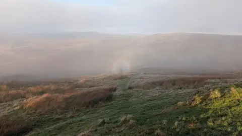 Steve Cartwright A foggy hillside with green grass and long, brown grass in the foreground. A circular rainbow is at the centre of the image. In the middle appears to be the outline of a man in dark, shadowy tones. The sky above is grey with a small amount of blue shining through the clouds.