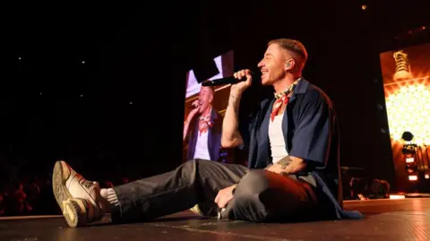 Getty Images Macklemore sits down on stage, smiling, holding a microphone up to his face. There is also an image of him on a screen behind him