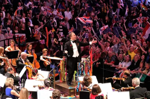 Conductor Sakari Oramo is pictured against a colourful backdrop of orchestral players and flag-waving audience members at the Last Night of the Proms in 2014