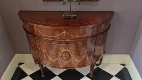 Pro Auction A demilune commode with a top inlaid with satinwood and ebonised strung banding, below are three drawers with conforming inlay together with bell flower swags, drapery and an oval fan medallion, on tapered square legs ending in brass capping.