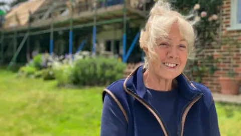 Angie Langley is smiling at the camera. She has blonde hair and is wearing a navy top with a navy fleeced gillet. She is sat infrontof a thatched cottage which has scaffolding on it. 