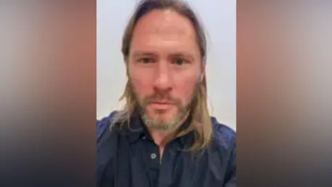 Justin Rundle looks towards the camera. He has long brown hair and a beard. He is wearing a blue shit. 