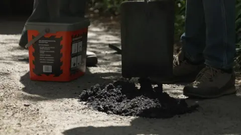 A pothole being repaired. A black tarmac-like product has been piled into a hole, ready to be pressed down.
