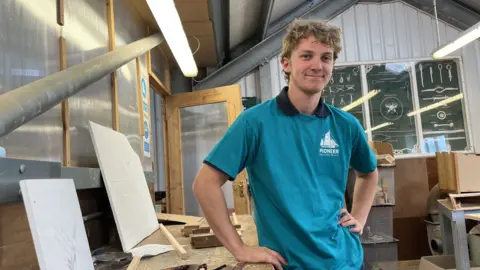 Martin Giles/BBC A young man stands in a workshop, surrounded by pieces of wood. He has his left hand on his hip and his right hand on the workbench. He is smiling at the camera. He is wearing a turquoise T-shirt with the Pioneer Sailing Trust logo on the left breast and a navy blue polo shirt underneath.