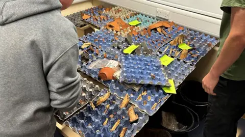 RAU Cadets looking at the findings. The artefacts are spread out on a table and neon post-it notes can be seen next to some of them. 
