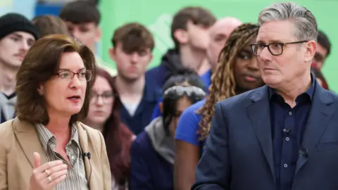 PA Media Eluned Morgan, a woman with brown hair and glasses, speaks in front of a crowd of people while standing next to Sir Keir Starmer, a man with grey hair and glasses.