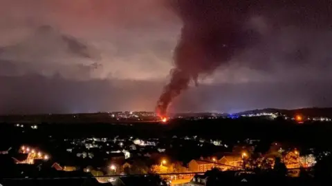 Adam Tilt A fire seen in the background with smoke pouring out into the sky. There are houses light by lamp posts in the foreground.