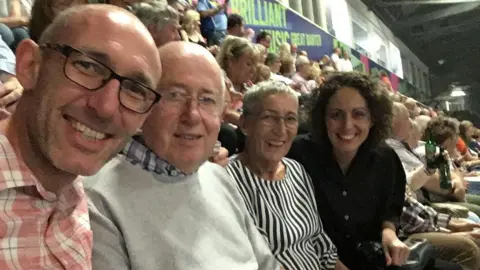 Family photo Two men and two women are smiling as they look at the camera while sitting in a row in a stadium, with a crowd of people around them.