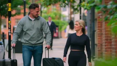 Netflix Jed and Bardha walking along the pavement, with Jed wheeling two suitcases