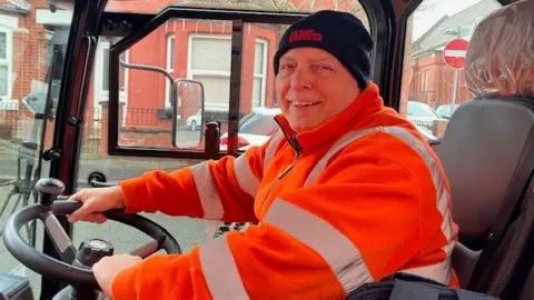 Ricky, a man in his 60s, sits in a clear-sided road sweeping vehicle. He wears an orange high-visibility jacket and a black woolly hat. He is smiling.
