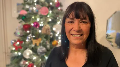 A woman with shoulder length dark hair, wearing a black top standing in front of a Christmas tree.