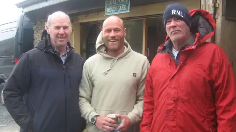 David Lewington (left) RNLI Supervisor Martyn Ward (right) and his son Joshua smile at the camera in coats and hoodies while stood in front of the Chapel Porth Beach Cafe 
