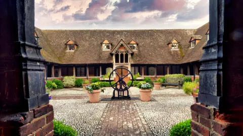 The almshouses are an old building with four sides and a cobbled central area. They have a steeply pitched roof with ornate dormer windows and a covered walkway around the bottom with white doors leading off from it. The central court is nearly planted with benches in the corner.